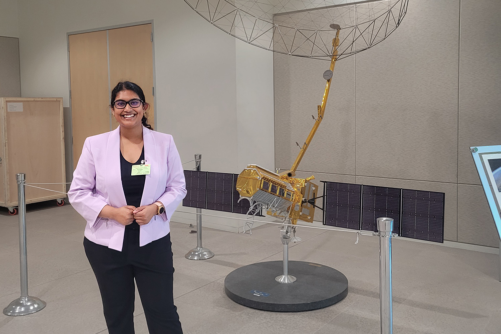Sreya Kumpatla stands next to a 1/6th scale model of the NISAR spacecraft.