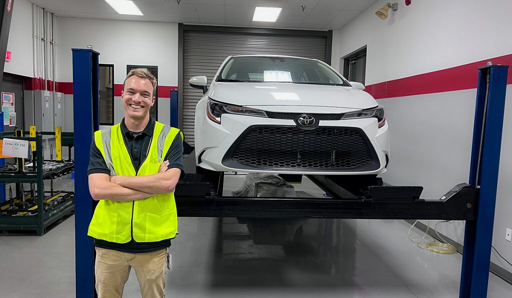 Carson Foy wears a bright yellow high-visibility vest as he stands in front a white Toyota that is raised on a rack in a garage.